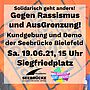 Im Hintergrund Schriftzug "Kein mensch ist illegal"; im Vordergrund Schirftzug "Solidarisch geht anders!" "gegen Rassismus und AusGrenzung. Kundgebung und Demo der Seebrücke Biefeld"